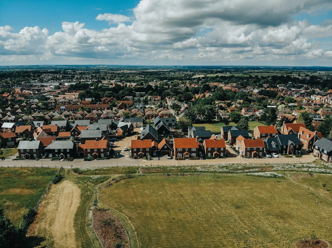 Aerial view of a suburban neighborhood with rows of houses, green fields, and a road under a partly cloudy sky.