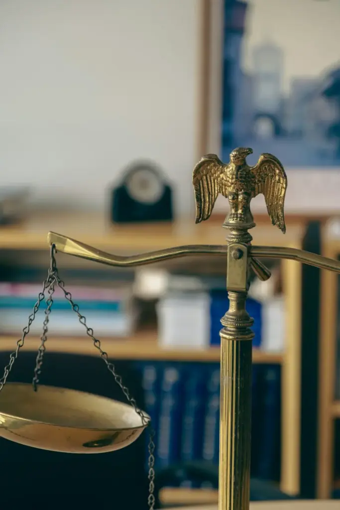 Close-up of a brass balance scale topped with an eagle finial, with blurred law books on shelves behind it.
