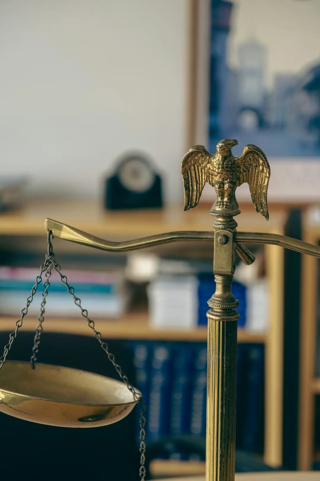 Close-up of a brass balance scale topped with an eagle finial, with blurred law books on shelves behind it.