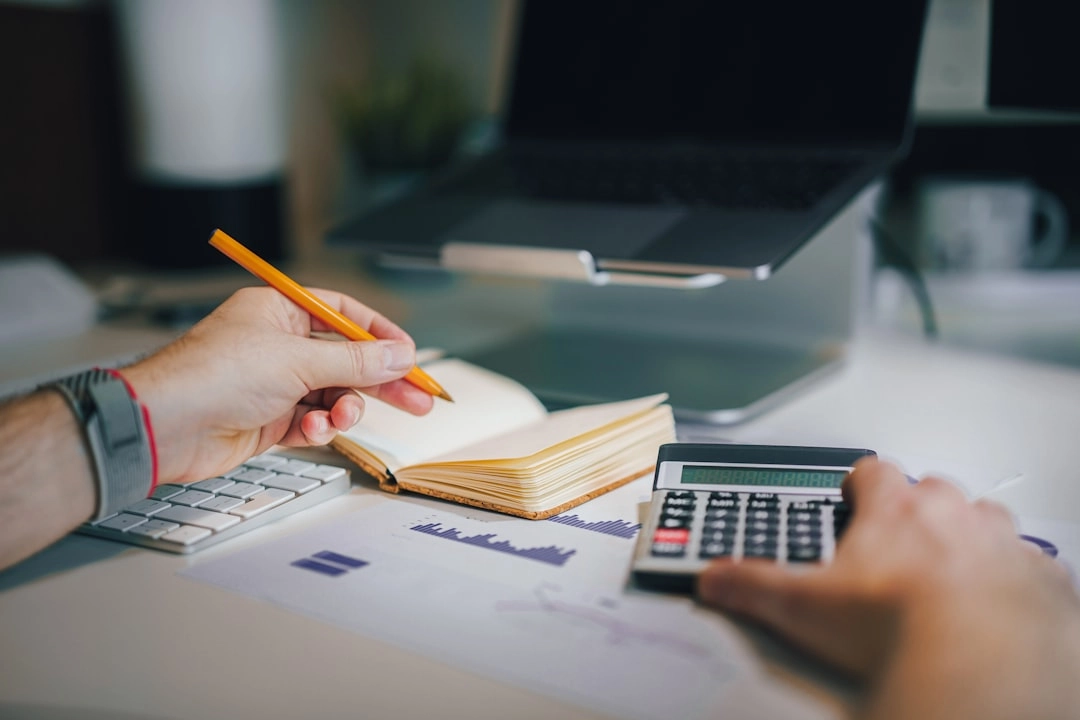 Hands using a calculator and holding a pencil over a notebook and financial charts at a desk with a laptop.