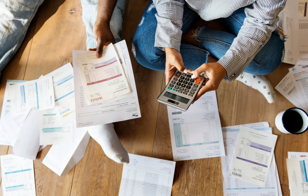 Two people sitting on a wood floor surrounded by invoices, one holding a calculator and the other holding paperwork.