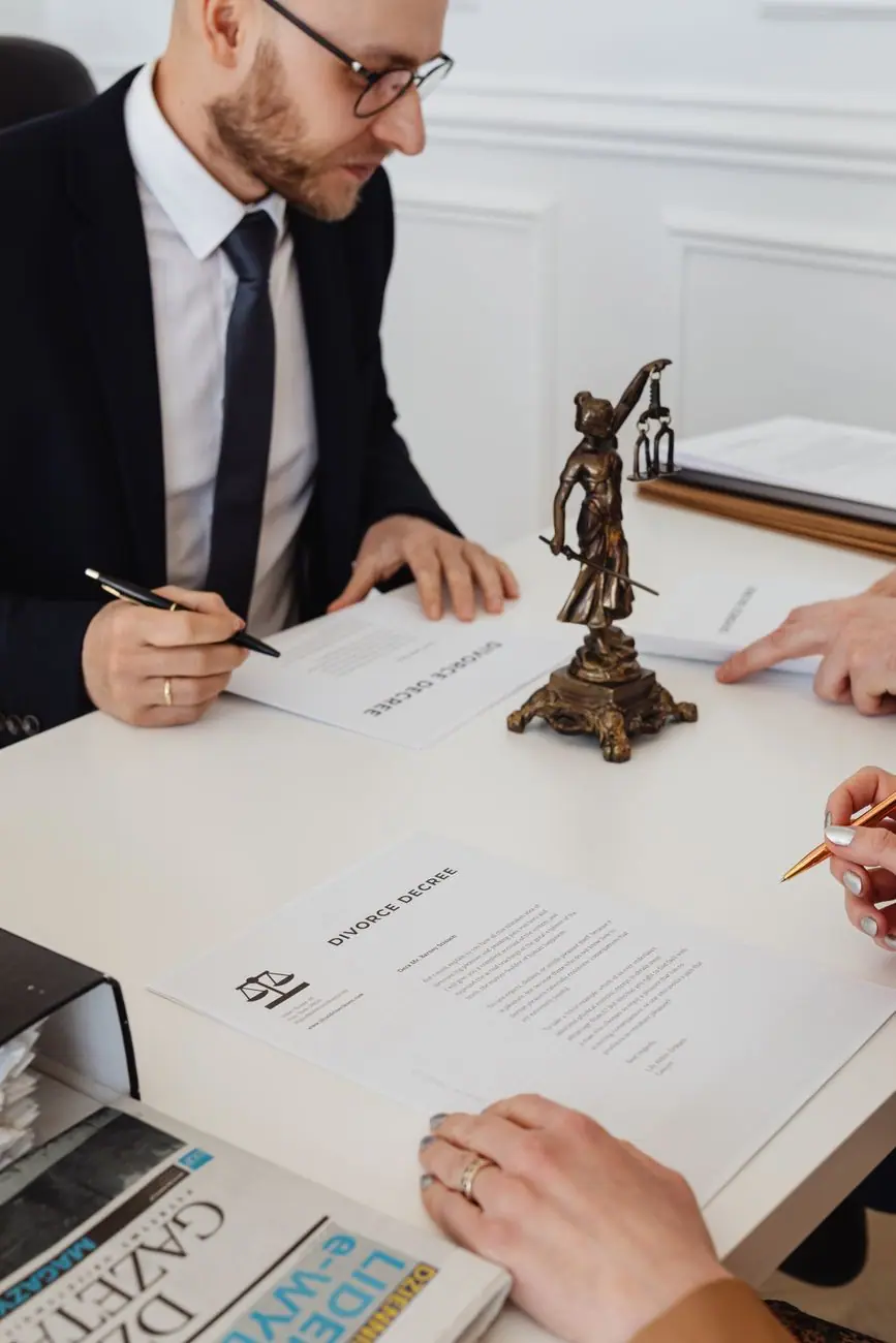 Attorney in a suit and two clients reviewing divorce decree papers at a white desk with a bronze Lady Justice statue.