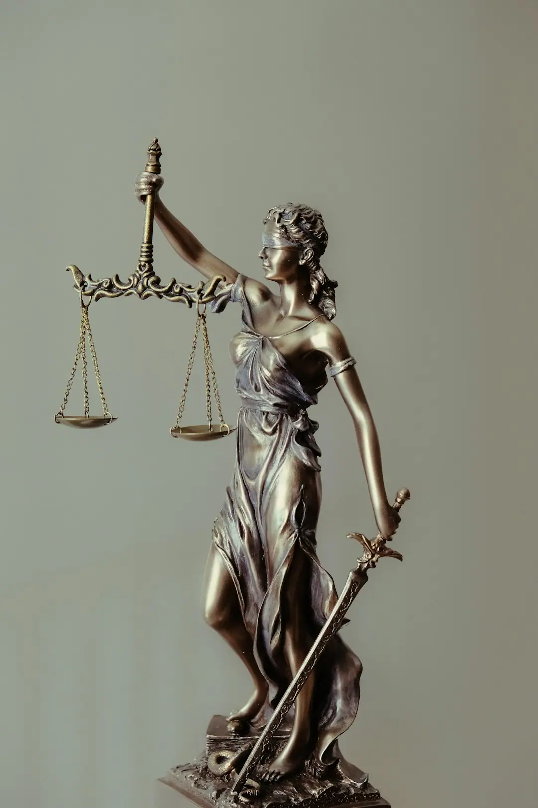 Bronze Lady Justice statue holding scales and a sword, blindfolded, against a plain gray background.