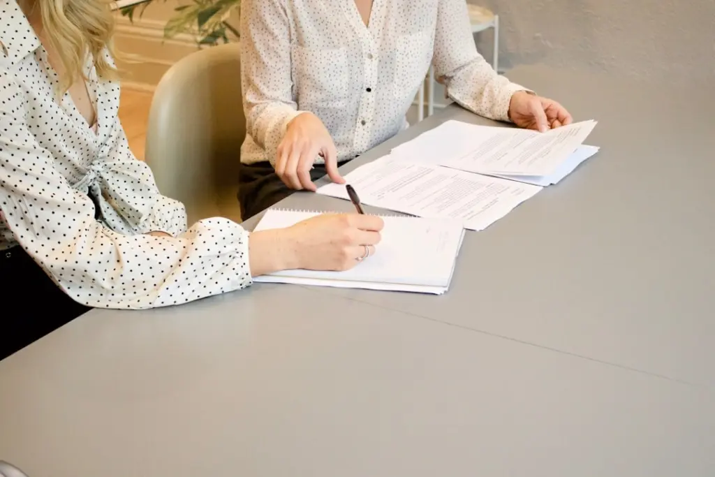 Two women at a gray table reviewing paperwork, one taking notes in a notebook while the other holds documents.