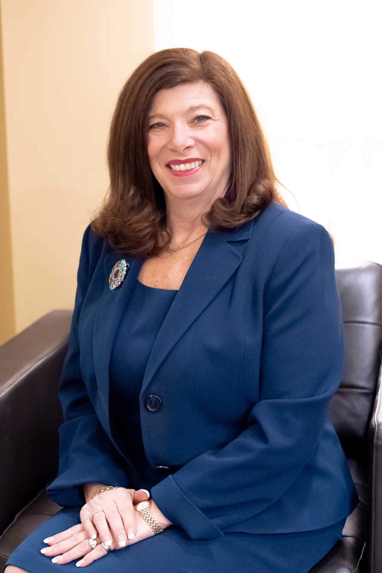 Smiling woman with shoulder-length brown hair wearing a navy blue suit and brooch, seated in a black leather chair.