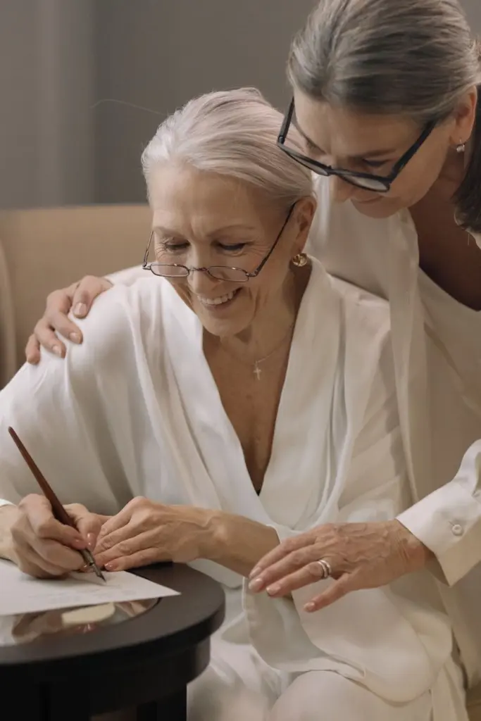 Smiling senior woman signing a document with a pen while another woman leans over her shoulder to assist.