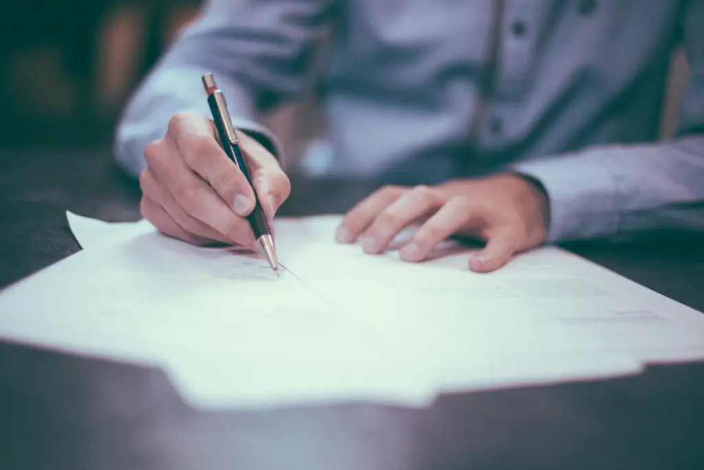 Close-up of a man in a blue shirt signing paperwork with a pen at a desk